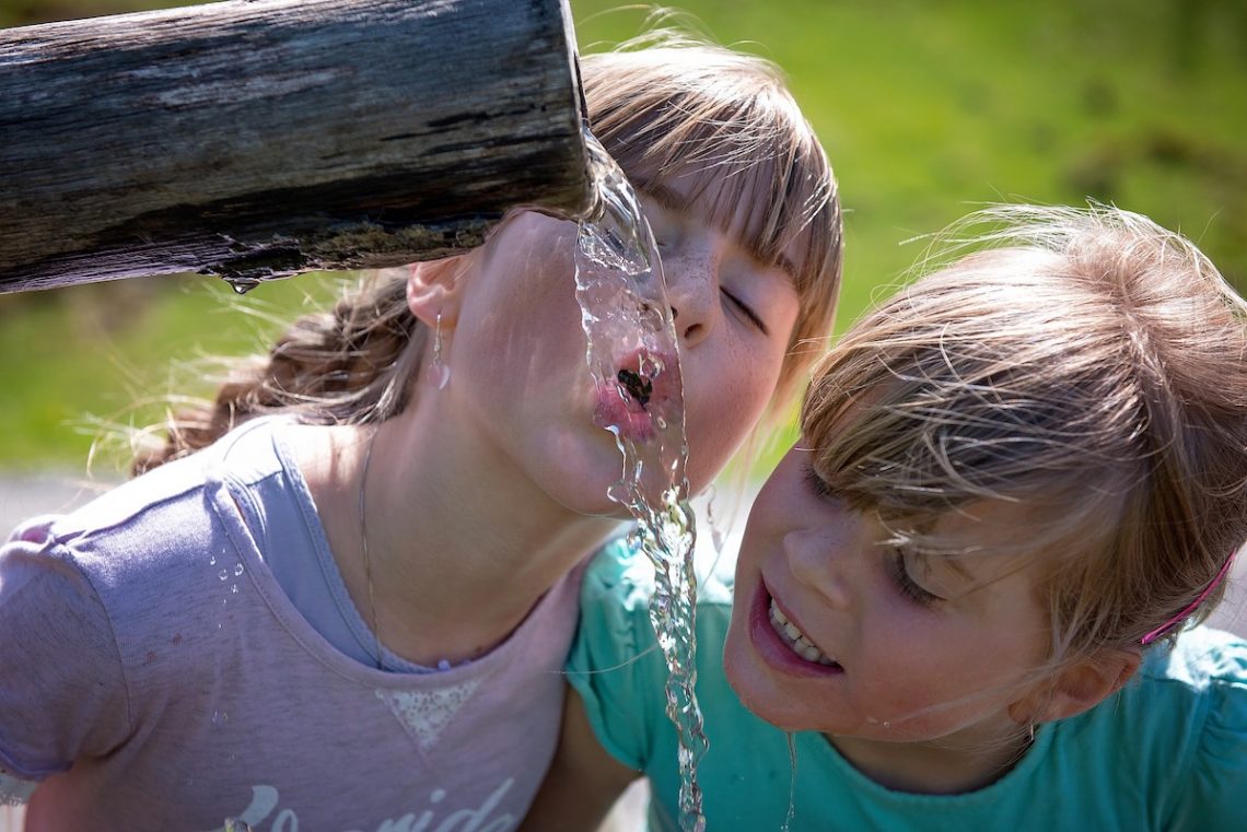 2 white young girls drinking clear water flowing from a pipe on a green blurred background.