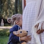 little girl hiding in woman's white skirt holding a stuffed animal